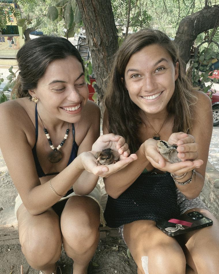 Dos mujeres riendo, sentadas a la sombra de un árbol, sosteniendo dos pollitos en las manos